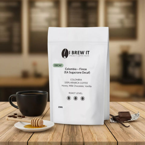 Colombia Fincas Mustafa (EA Sugarcane Decaf) 250g bag with brewed cup on wooden table, warm bokeh, honey dipper, vanilla pod and milk chocolate props, DECAF.