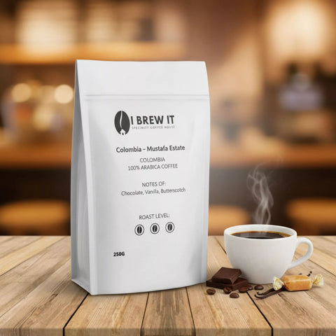 Colombia Mustafa Estate 250g bag with brewed cup on wooden table, warm bokeh, chocolate square, vanilla pod and butterscotch piece props.