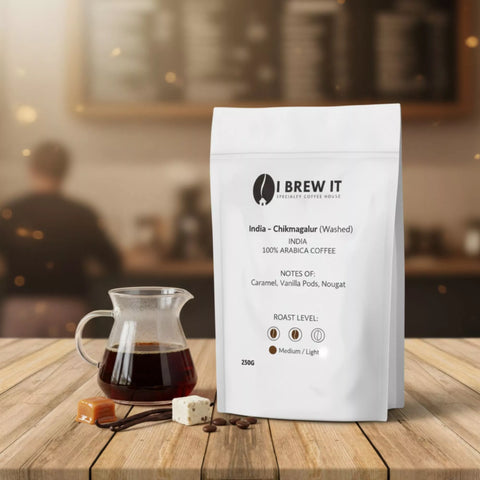 India Chikmagalur (Washed) 250g bag with brewed cup on wooden table, warm coffee-shop bokeh, caramel, vanilla pod and nougat props.