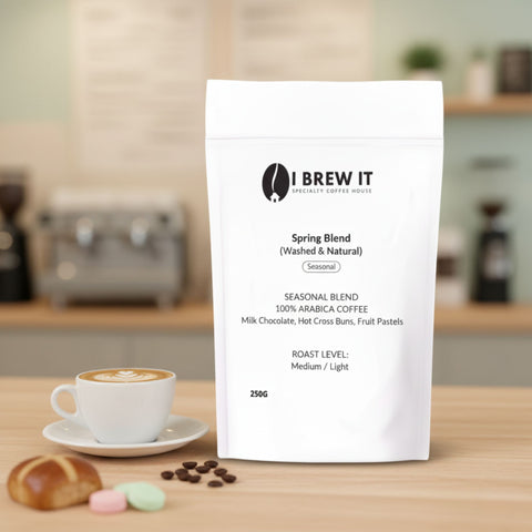 Spring Blend 250g bag with brewed cup on wooden table, warm spring bokeh, mini hot cross bun and pastel sweets props.